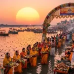 चैत्र छठ पूजा 2026: पटना, बिहार के गंगा घाट पर सूर्योदय के समय व्रती और उनके प्रसाद के सूप। फूलों के तोरण द्वार पर कार्यक्रम का बैनर स्पष्ट है।